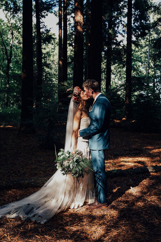 Boho bride in a strapless wedding dress and flower crown kissing her groom in the woods with the sun shining through the trees 