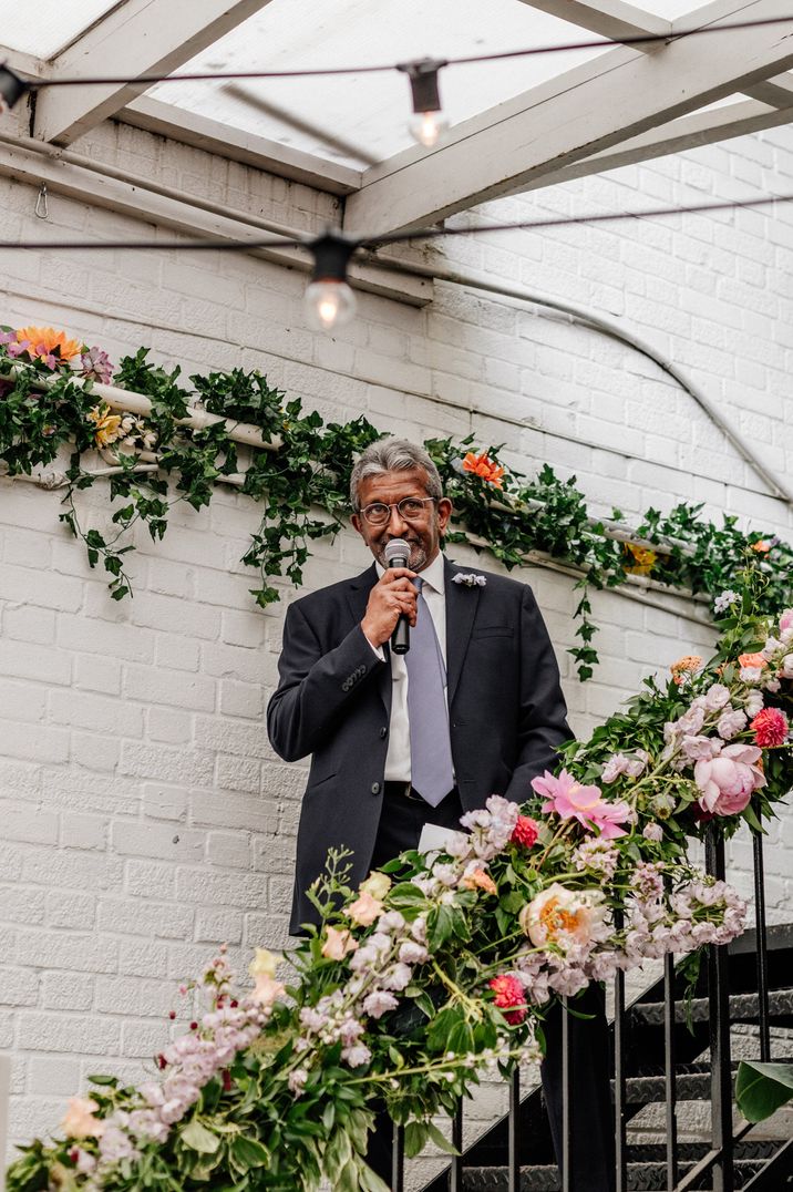 Person making wedding speech in black suit and grey tie on floral staircase 