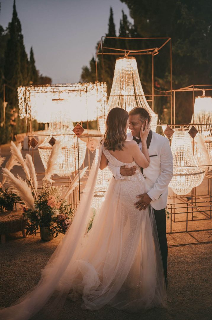 The bride smiles with the groom in a black and white wedding tux next to the chandelier wedding table plan 