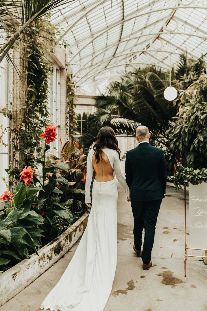 Bride in open back wedding dress with long sleeves and groom in black suit walking through glass house wedding venues Syon Park with tropical flowers and large palm trees 