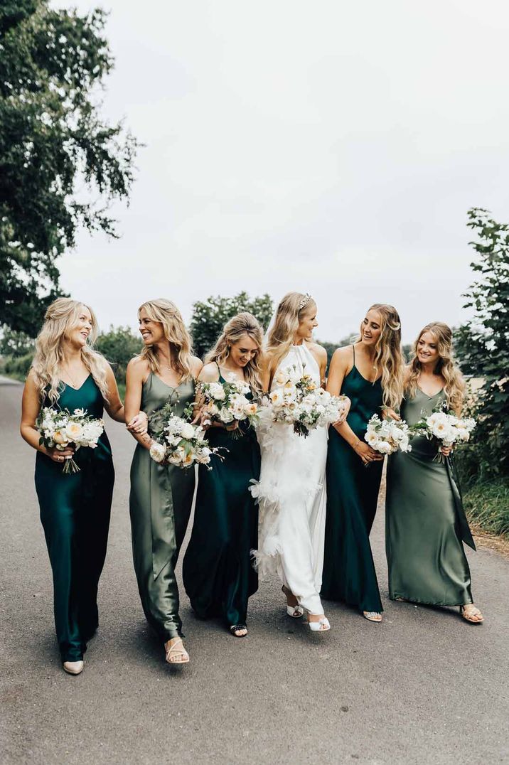 Bride in halterneck wedding dress walking with bridesmaids in mismatched green satin bridesmaid dresses 