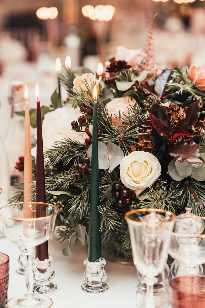 Dark green, burgundy and cream taper candles with festive flower arrangements as the table centrepiece with winter berries and more 