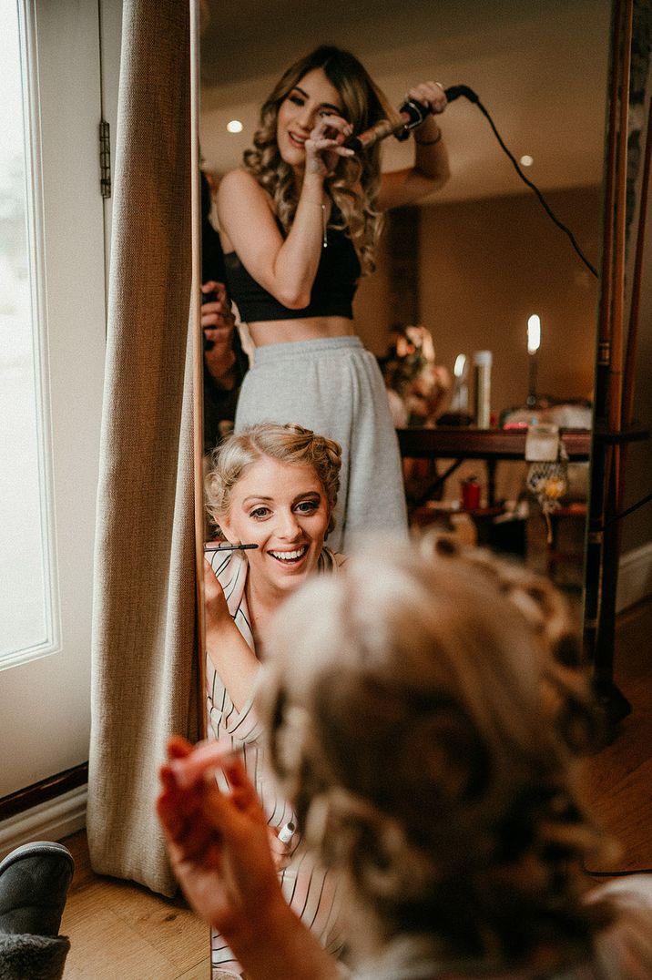 Two girls get ready together facing a mirror as one of them does their hair and another does her makeup 