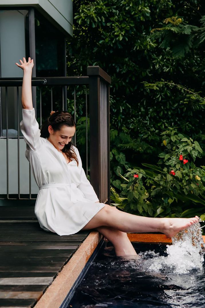 Bride enjoying hot tub pool by dipping her feet in wearing a white robe by Emma-Jane Photography