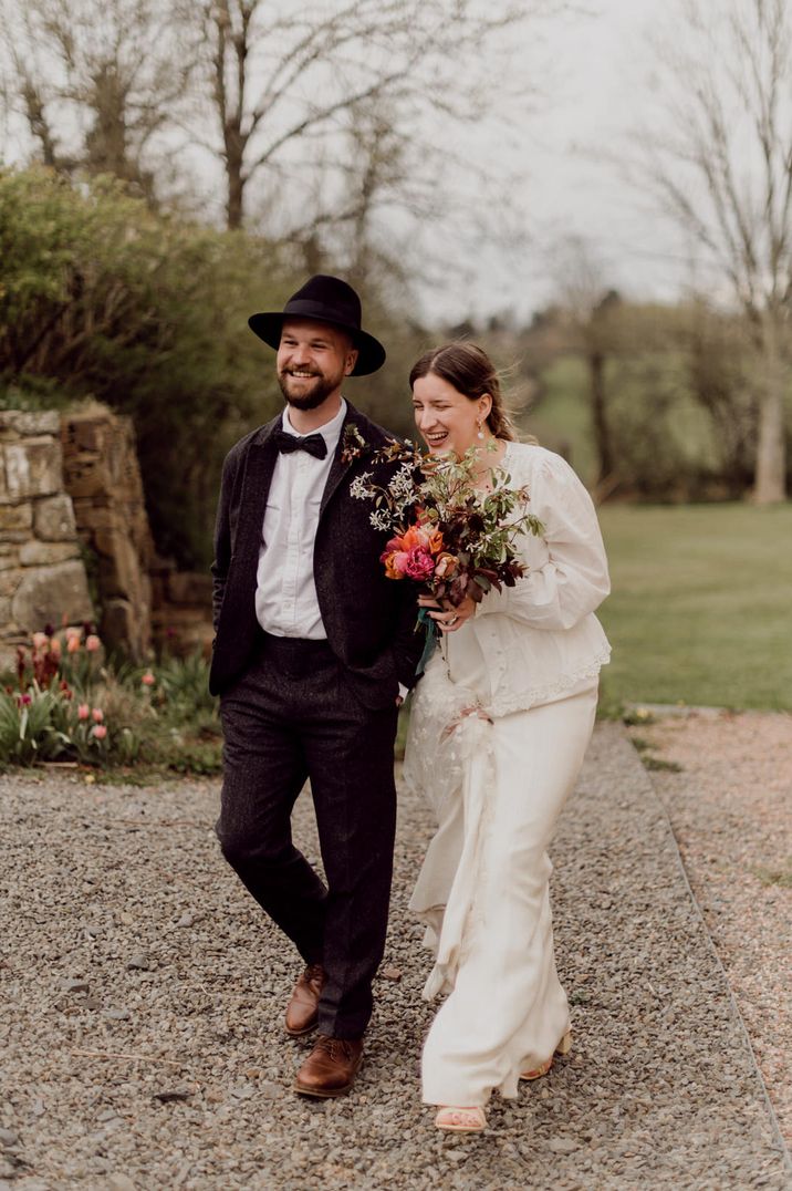 Groom in a black suit bow tie and fedora hat arm in arm with his bride in an appliqué veil holding a red bouquet 