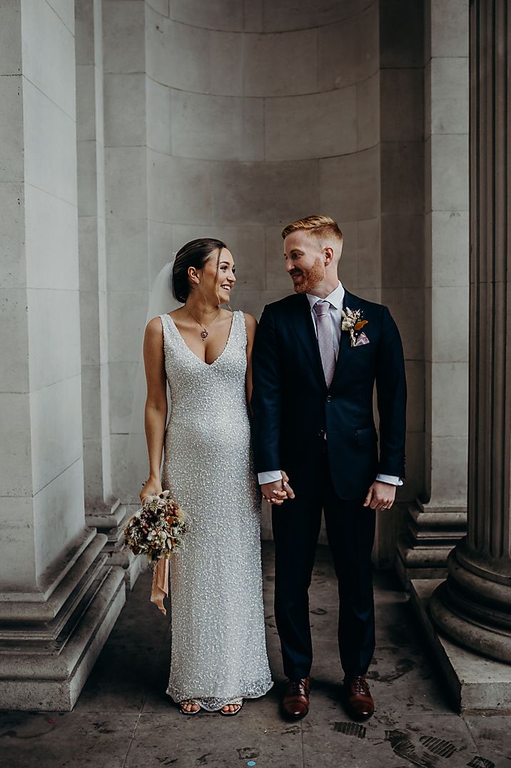 Groom in blue suit with a purple tie standing holding hands with the bride in a sparkly ASOS wedding dress 