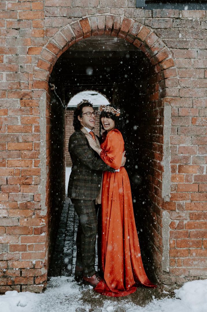 Groom in a brown checkered suit with the bride in a red long sleeve wedding dress hide from the snow