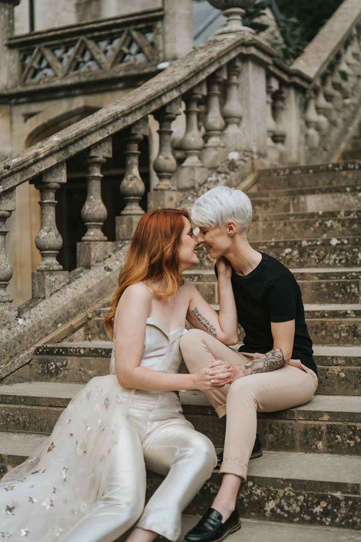 Bride in golden sheer bridal jumpsuit with sheer gold fleck train attached at the hip embracing bride on stairs at LGBTQIA+ wedding