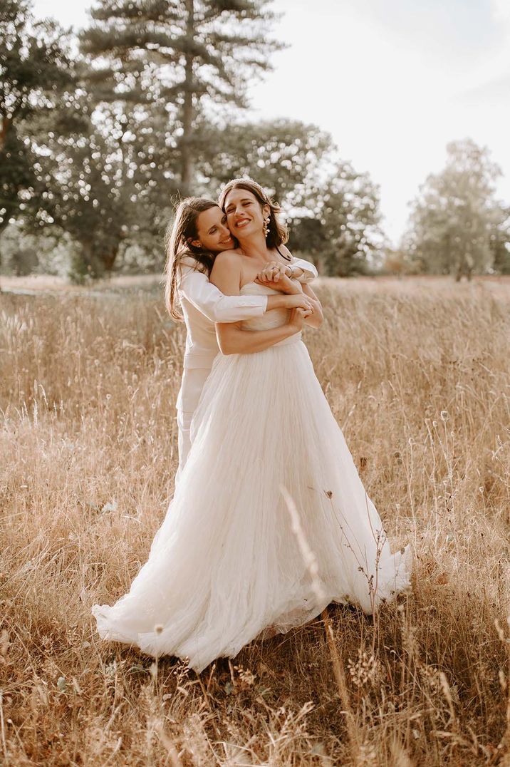 Bride in strapless wedding dress being embraced by the bride in a white pant suit for the outdoor wedding over 60k 