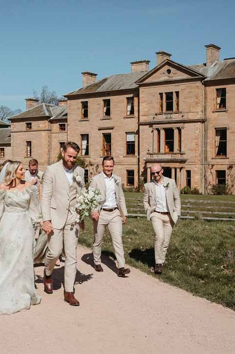 Bride in sage green off shoulder long sleeve wedding dress with groom in light suit with wedding party in sage green wedding outfits walking the grounds in one of the best wedding venues Scotland - Cambo House 