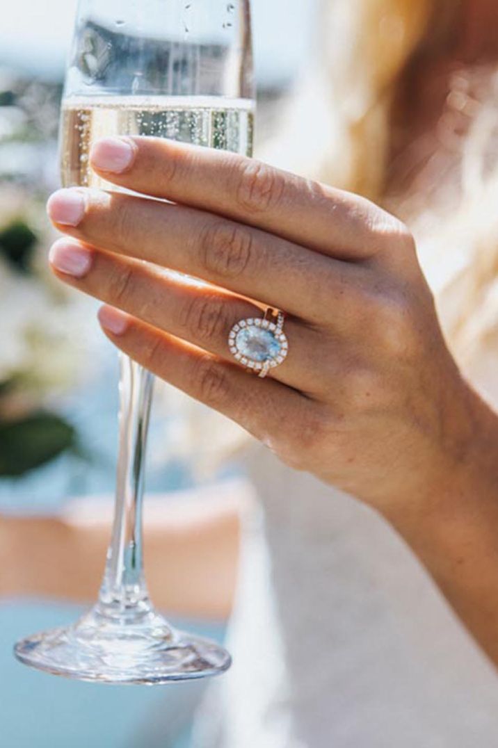 Light sapphire blue oval wedding ring with halo worn by woman drinking champagne 