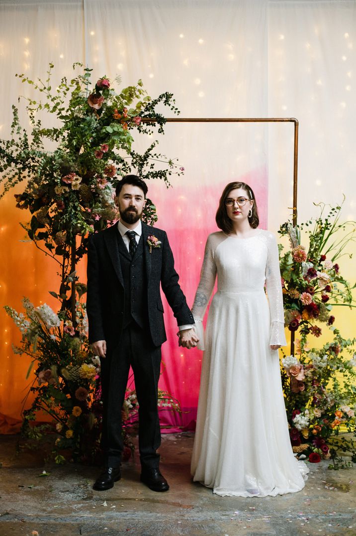 Bride in glasses and a sparkly Rolling In Roses wedding dress holding hands with her groom in wool suit in front of a copper pipe archway decorated with foliage and flowers in front of a tie-dye fabric cloth