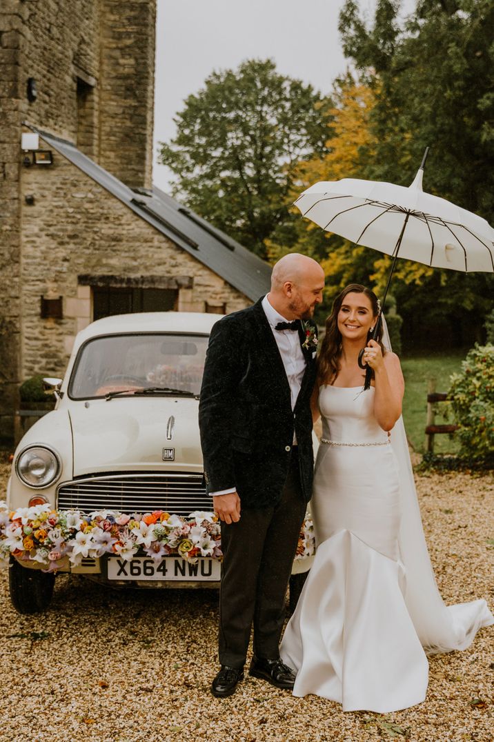 Bride in a strapless wedding dress and groom in a tuxedo holding an umbrella standing next to their Kushi Car wedding transport 