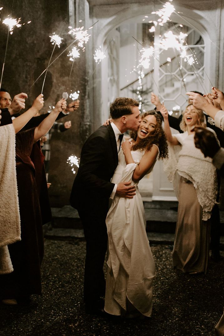 Groom in black tie kisses the bride on the cheek as she smiles widely and guests wave their sparklers 