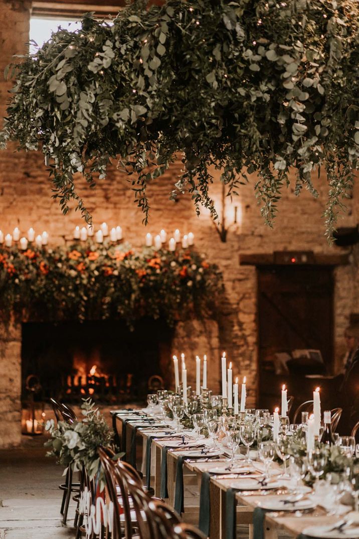 Rustic wedding tablescape at Cripps Barn wedding venue with suspended eucalyptus decor, white tapered candles, eucalyptus table runners and a roaring fireplace 