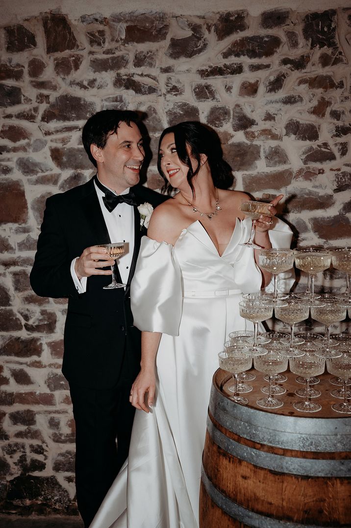 Champagne tower with the bride in a strapless wedding dress with detachable sleeves standing with the groom in a black tuxedo