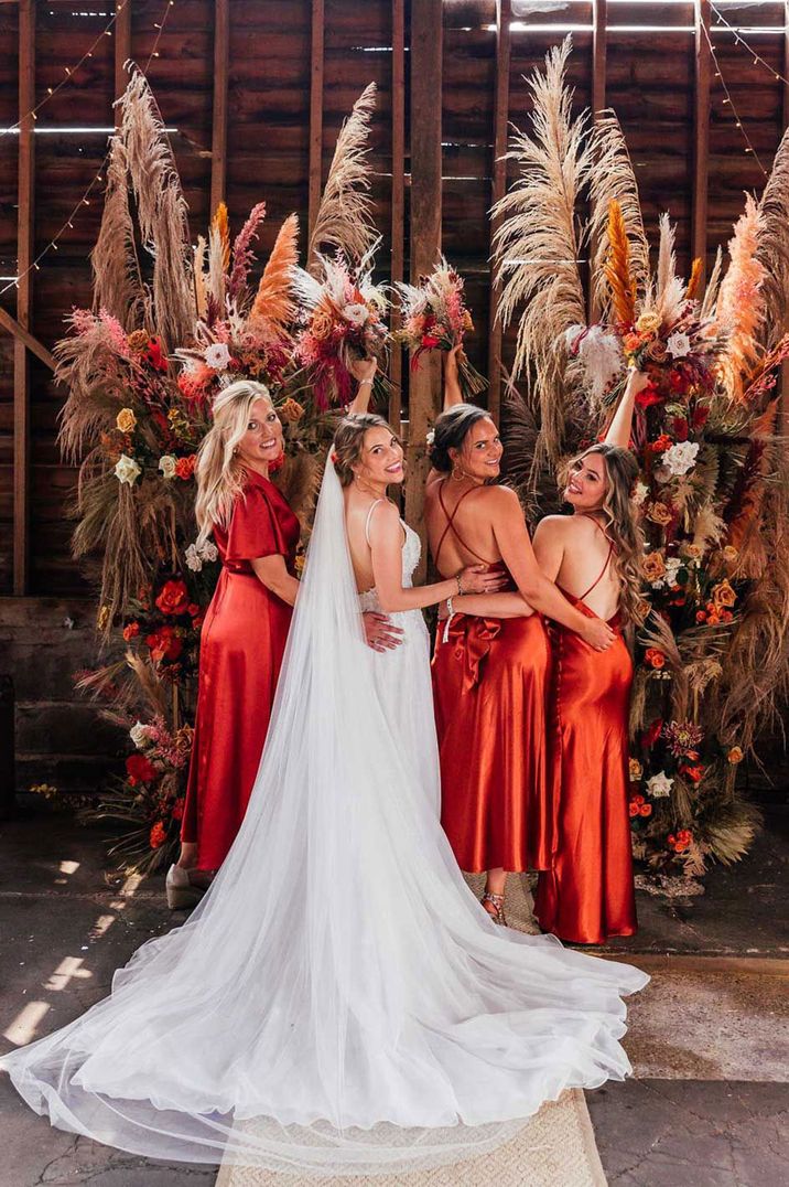 Bride in corset back wedding dress with train stands with bridesmaids in burnt orange satin bridesmaid dresses all holding up bouquets