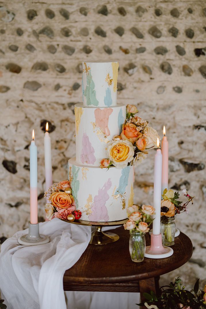 Three tier white iced wedding cake with pastel pink, blue, green, purple and yellow painted icing with ombre candles