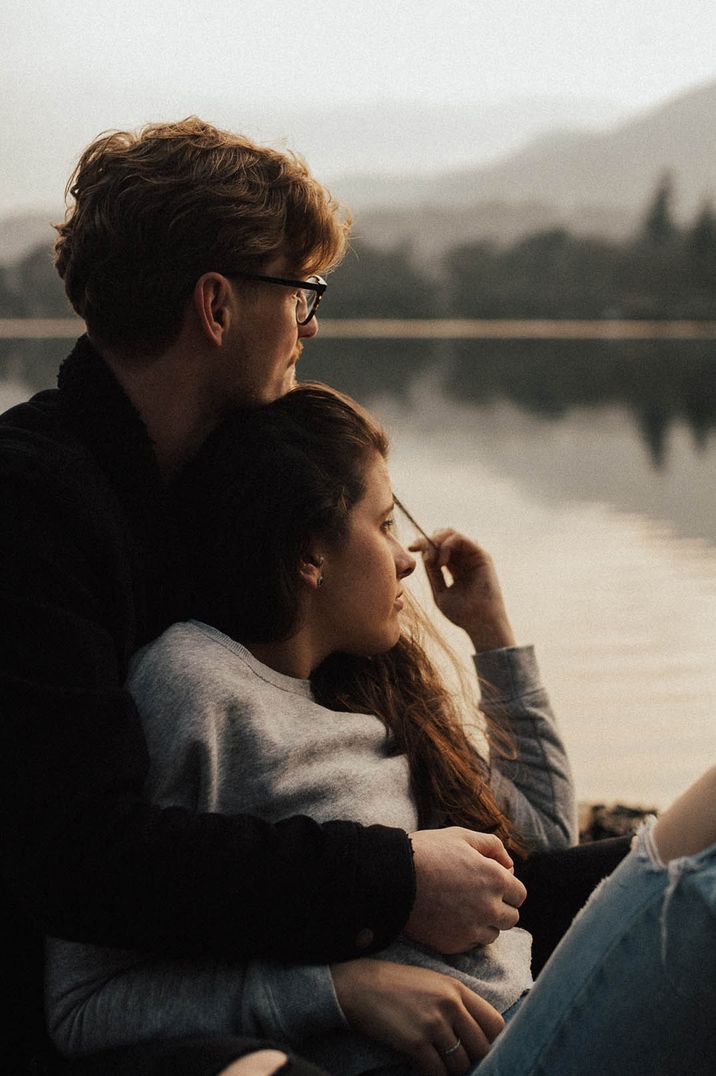 Engaged couple sit looking out over the view of a lake 