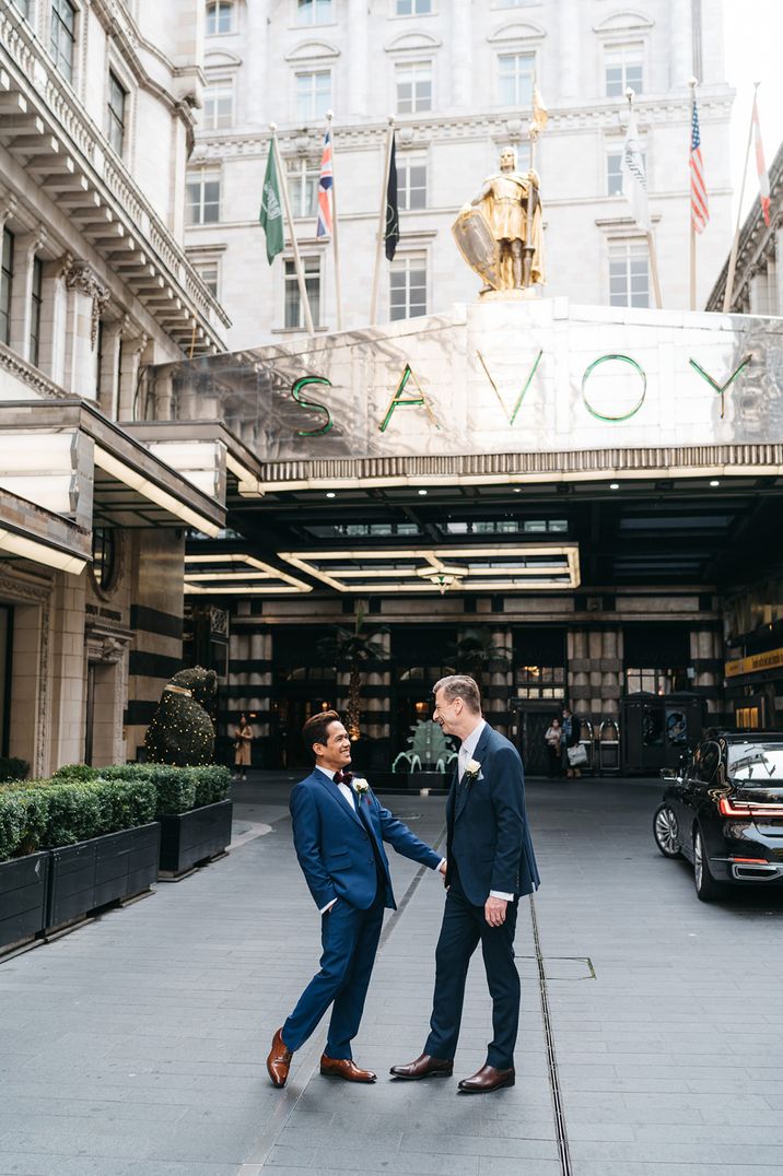 Grooms in blue suits holding hands outside the Savoy hotel at London elopement - how to save for your wedding