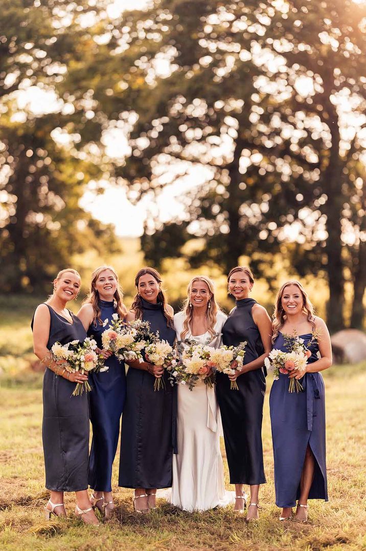 Bride in short sleeve satin wedding dress standing with bridal party in mismatched navy bridesmaid dresses 