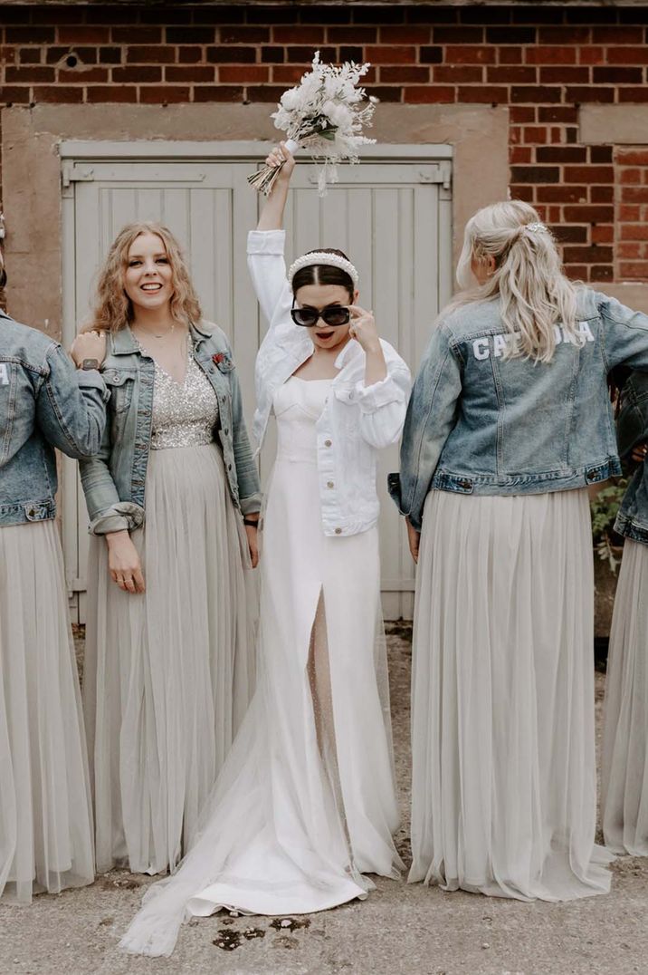 Bridesmaids in light blue denim jackets with their names on the back posing with the bride in a white bridal denim jacket and sunglasses 