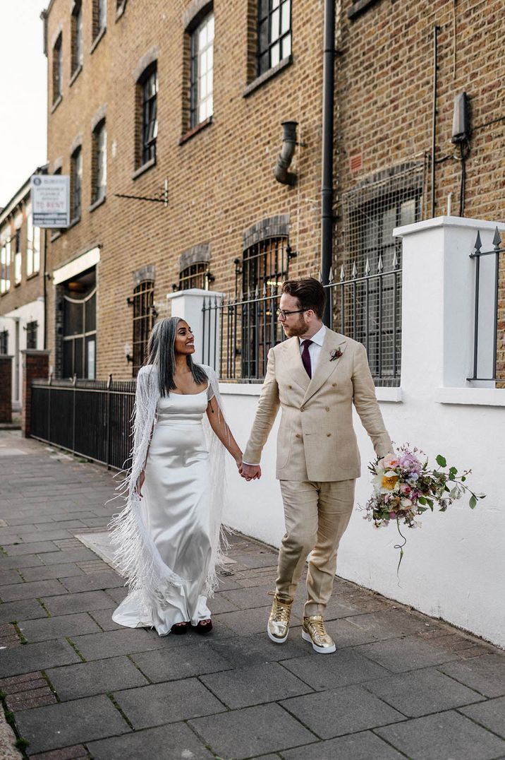 Bride in satin wedding dress with sparkly tassel bridal cape with the groom in a double breasted cream suit for city wedding 