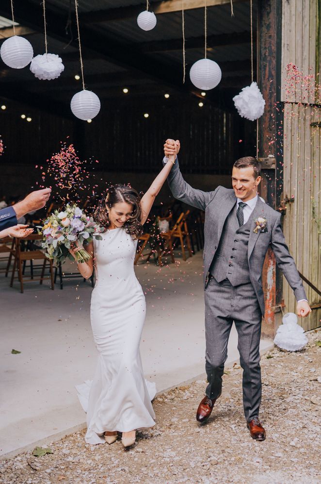 Bride and groom confetti exit out of a barn with white hanging lanterns and tissue balls decor