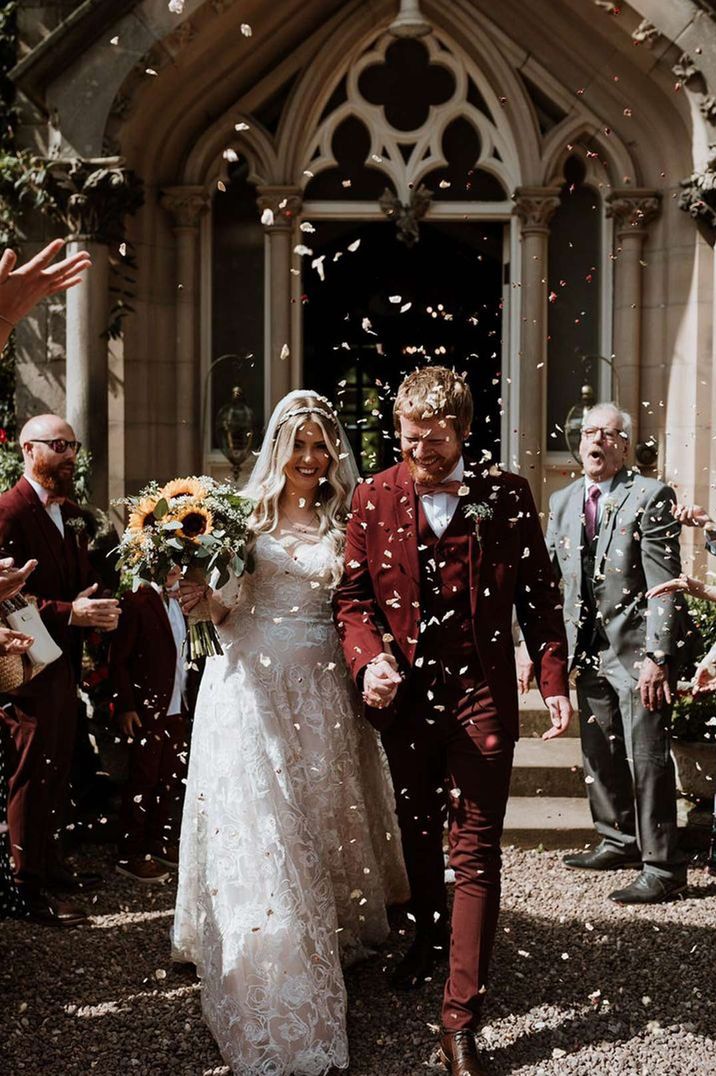 Groom in burgundy grooms suit holding hands with bride in lace wedding dress holding sunflower bridal bouquet doing confetti exit at Dunwood Hall Estate castle wedding venues 