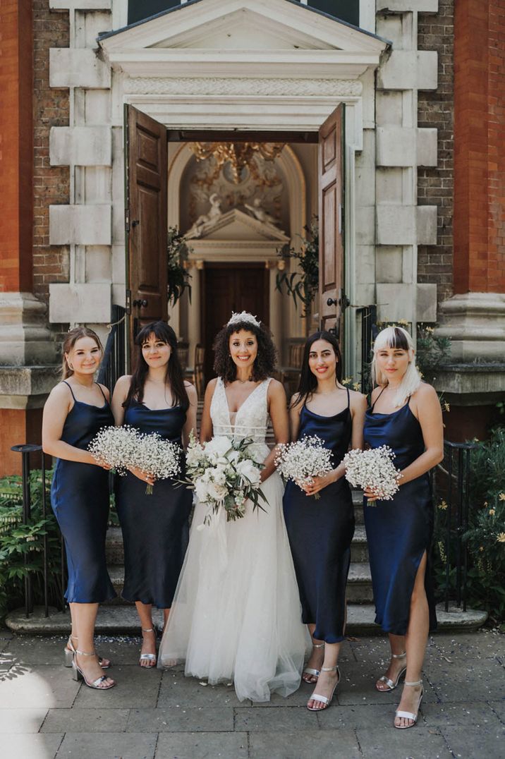 Bride in deep v neck sleeveless wedding dress standing with bridal party in strappy midi navy blue bridesmaid dresses 