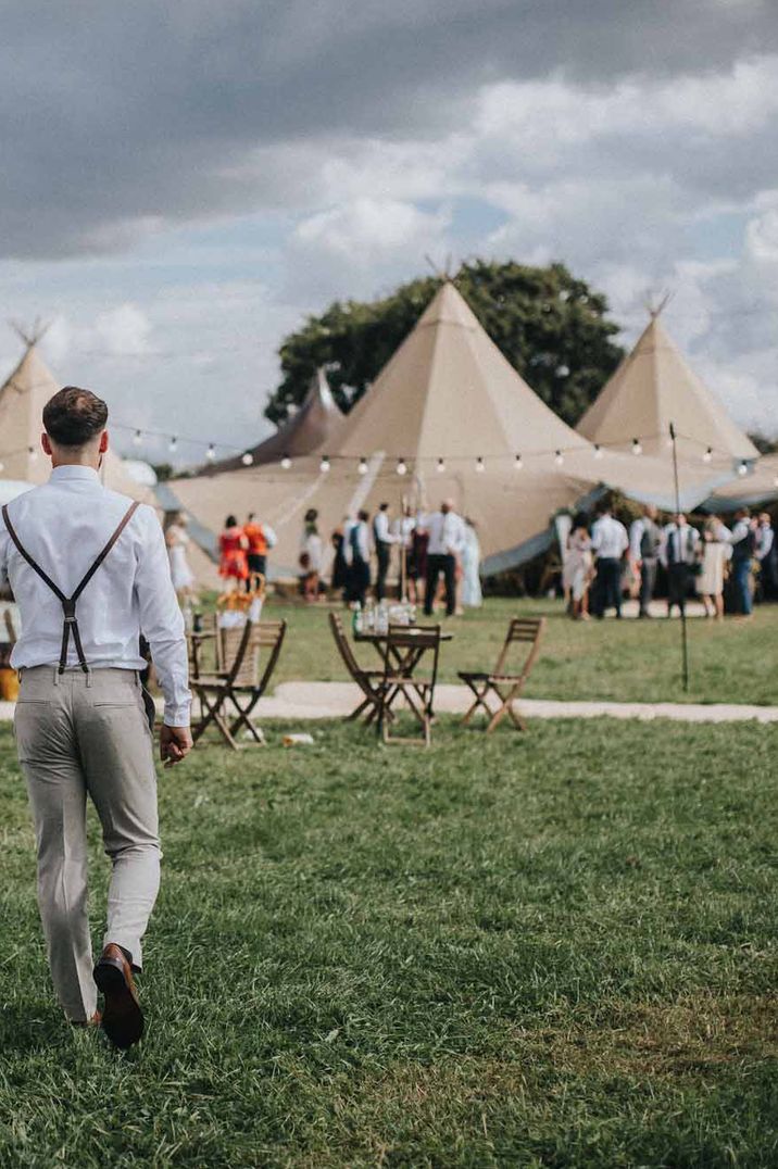 Bride and groom holding hands in a lace boho wedding dress and braces at their outdoor tipi wedding reception 