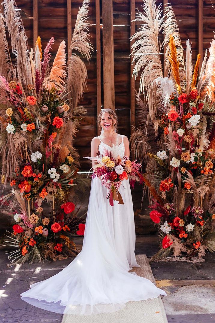 Bride in boho wedding dress holding an orange bouquet inbetween two pampas grass columns