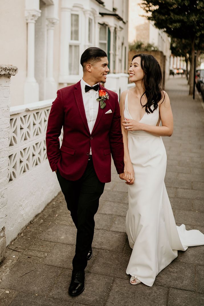 Groom in burgundy blazer and rose boutonniere walking with bride in v-neck satin wedding dress 