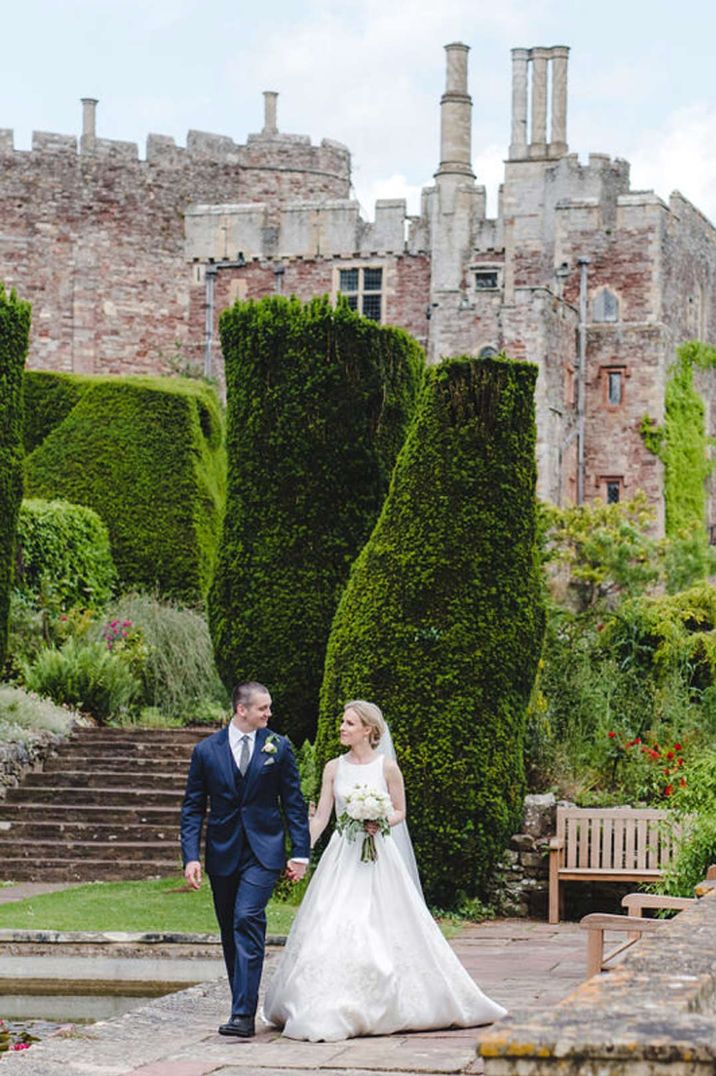 Bride in halterneck wedding dress and church length veil walking with groom in deep blue grooms suit on the grounds of Berkeley Castle wedding venue 