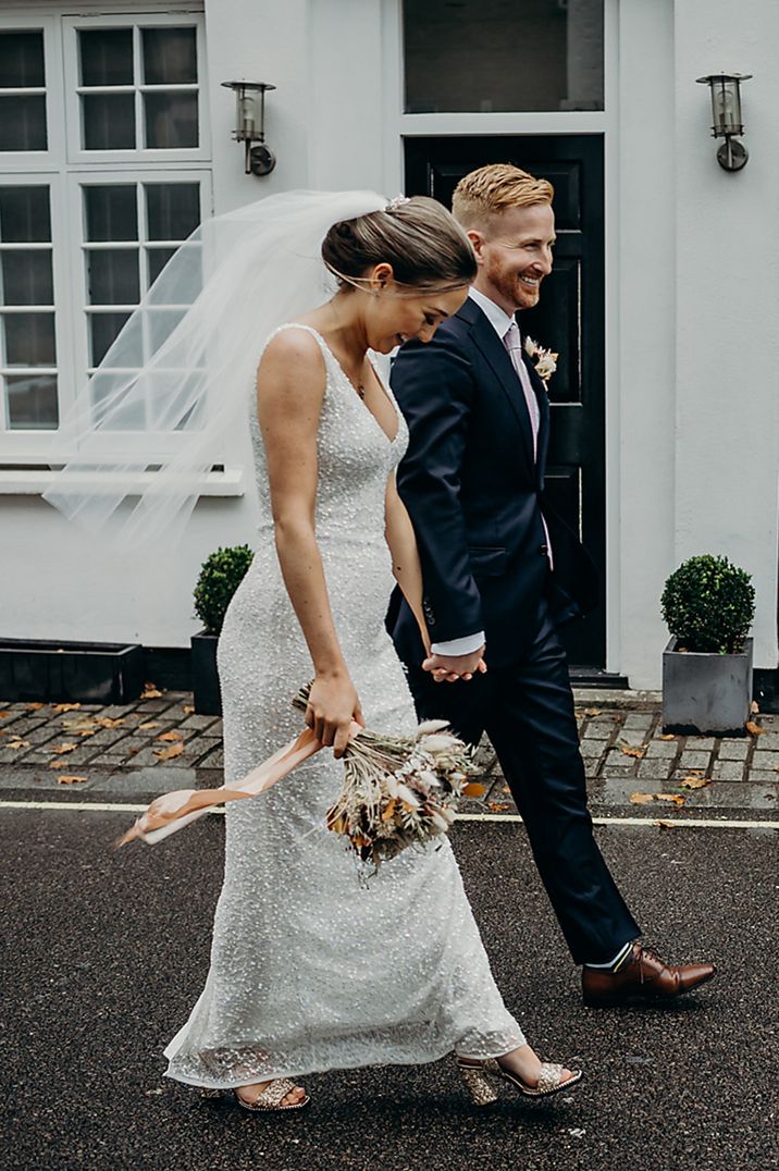 Groom in navy suit walking hand in hand with the bride in a sequin maternity wedding dress 