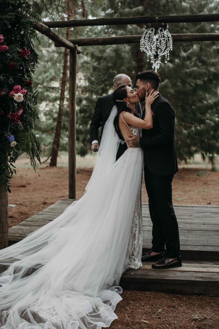 Bride and groom kiss outdoors in a wood under a chandelier