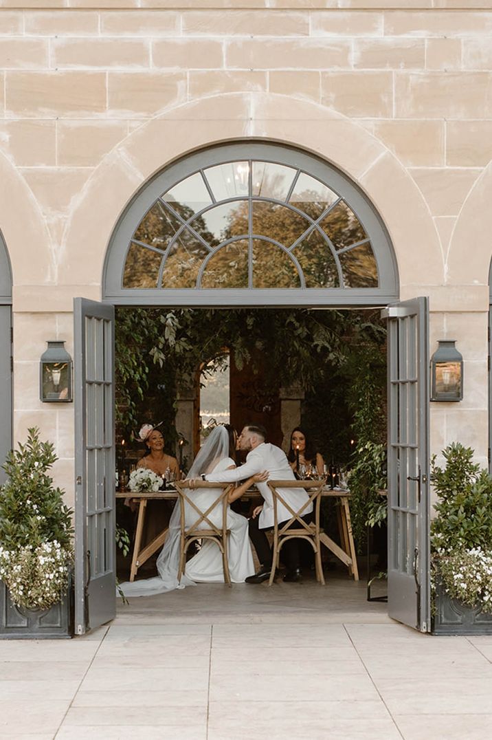 Bride and groom embrace, sat at their traditional wedding table through open doors