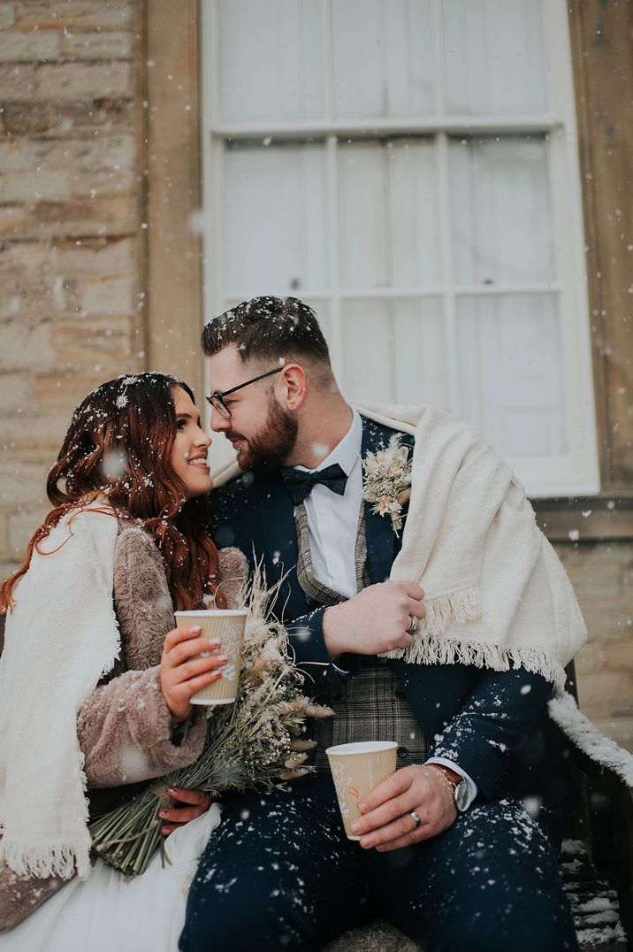 Bride and groom lean in close drinking hot drinks with a blanket on a snowy day 