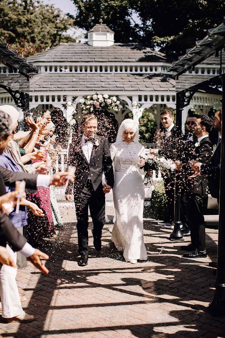 Book wedding venue - Bride in long sleeve lace wedding dress and groom in classic black tux doing confetti walk at outdoor summer wedding at The Old Kent Barn venue 