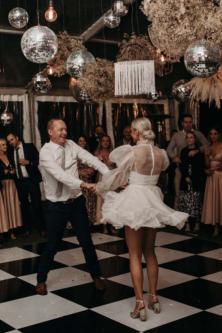 Bride in short wedding dress with sheer puff sleeves and back detail dancing with groom with hanging disco balls wedding decor and festoon lighting 