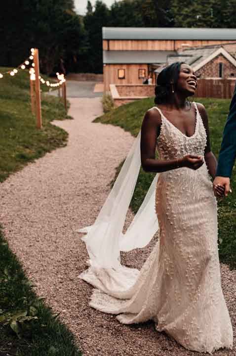 Bride in v neck strappy beaded wedding dress and Watteau train walking with groom in three piece deep teal suit - how much are wedding dresses