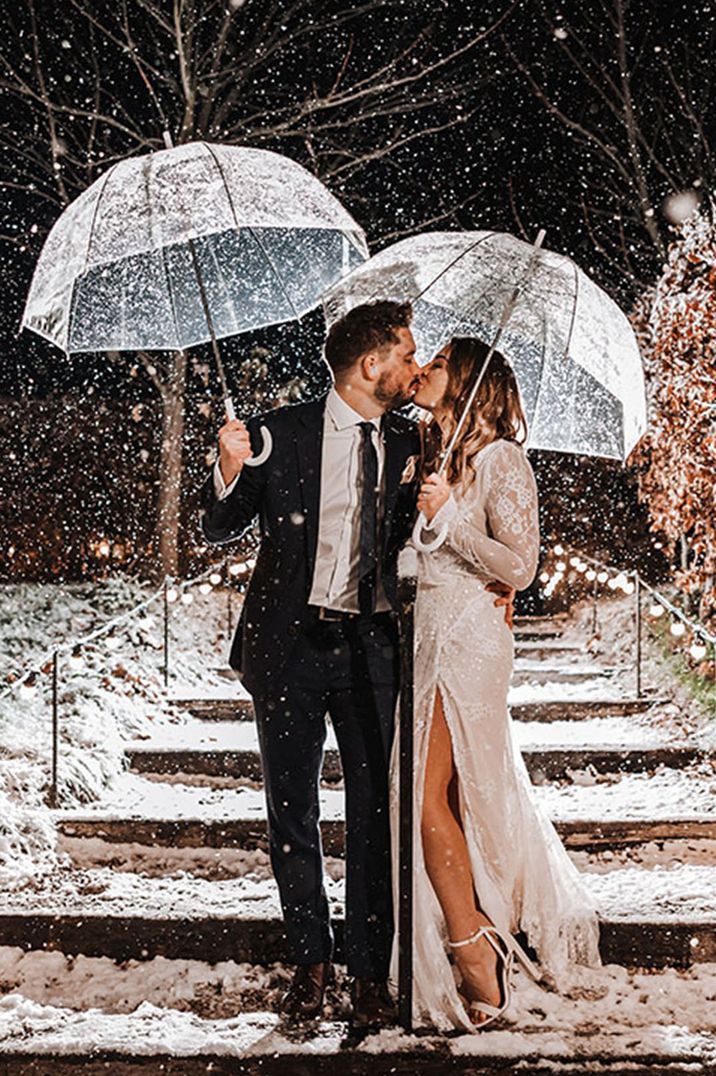 The bride and groom hold clear umbrellas at their snowy wedding  