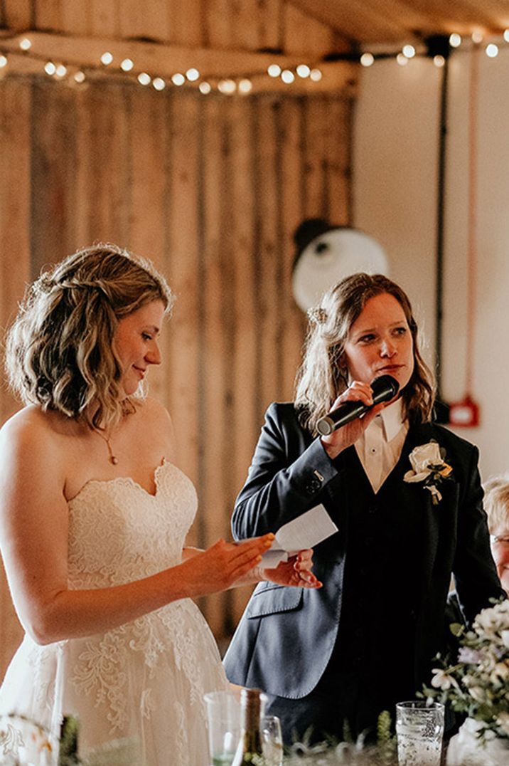 Bride in sweetheart lace wedding dress with bride in black suit with white rose buttonhole reading out their joint wedding speech 