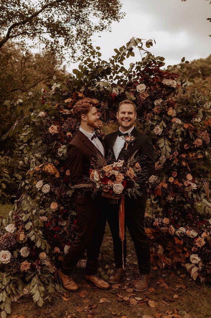 Two grooms standing in front of autumnal flower arch carrying autumnal bouquet with seasonal blooms 