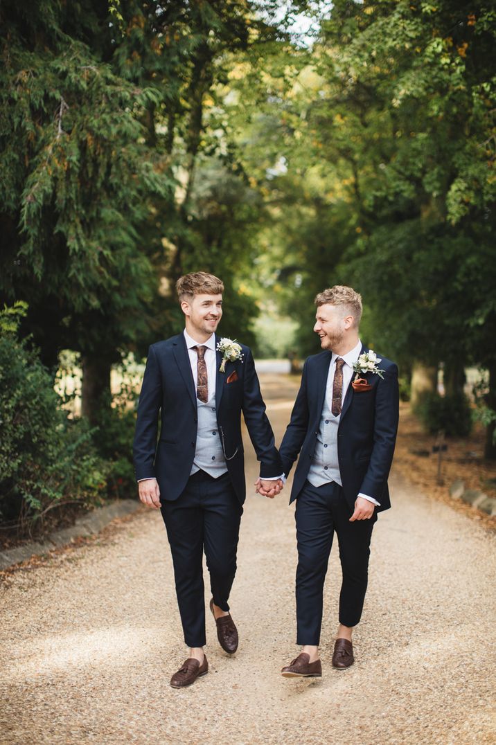 Two grooms walking in the greenery in matching dark blue suits 