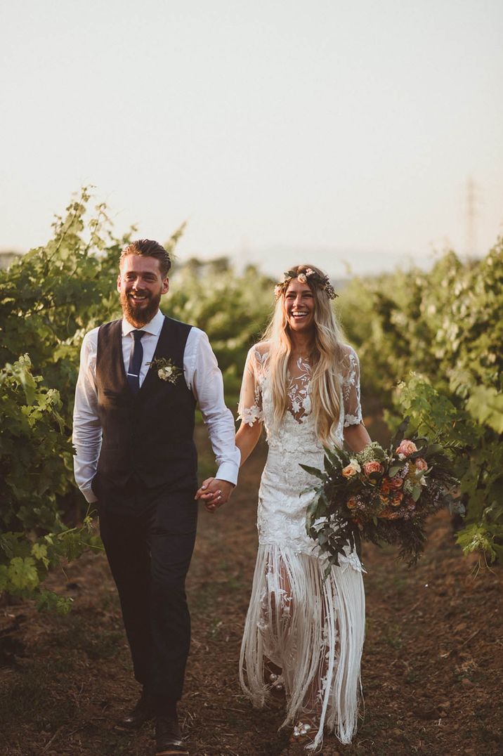 Groom in blue tie and black waistcoat and trousers walking with the bride in a boho lace handmade wedding dress with fringe 