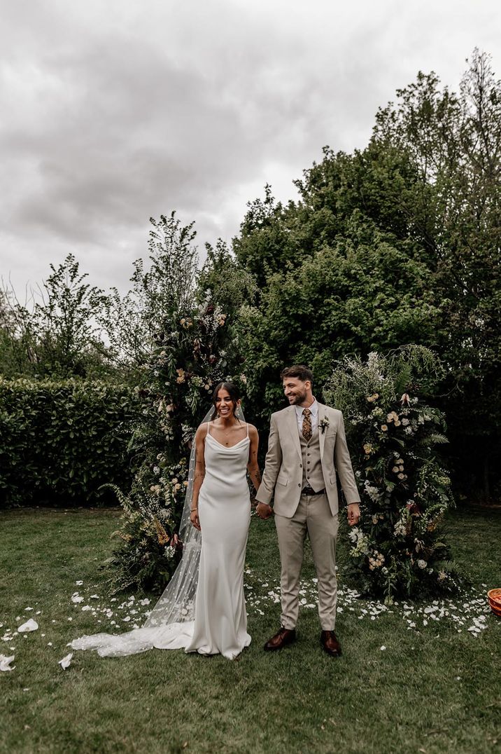 Outdoor back garden wedding over 60k with the bride in a satin wedding dress and groom in three piece cream suit 