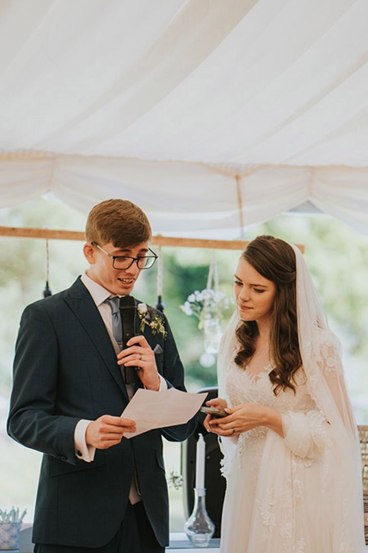 Groom in navy suit holding a microphone as he reads out a wedding speech with the bride standing next to him 