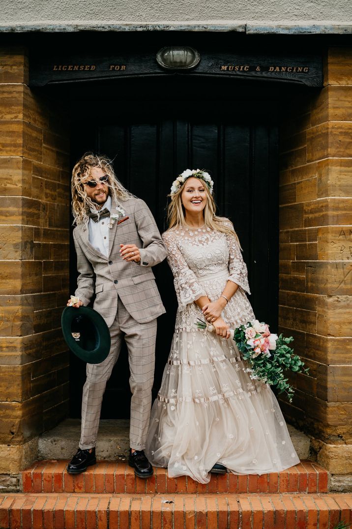 Bearded groom with long hair in a grey check suit and bow tie and bride with long hair wearing a flower crown and boho lace wedding dress