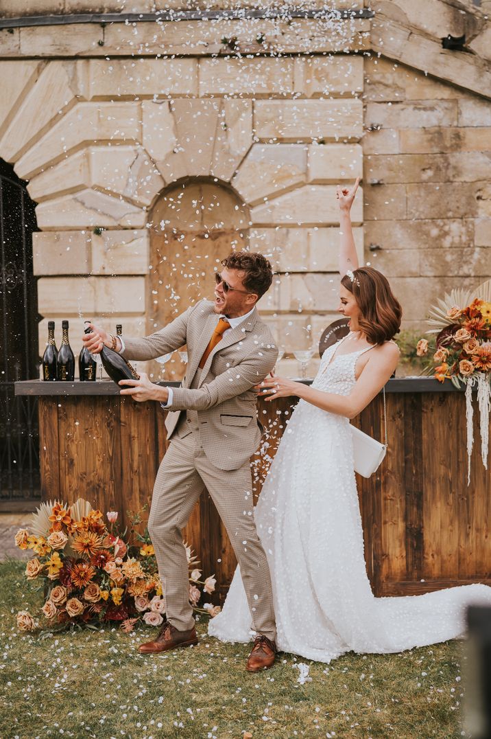 Couple in tartan suit and sleeveless lace wedding dress popping champagne with white confetti